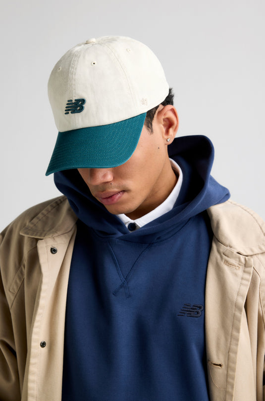 Man wearing New Balance by '47 hat with a teal peak and cream crown., blue New Balance hoodie and a brown over coat