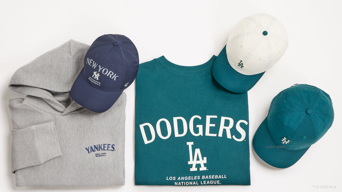 A flat-lay arrangement of baseball apparel on a white background: a folded gray hoodie with navy New York Yankees embroidery, a teal Los Angeles Dodgers T-shirt with large white DODGERS lettering and LA logo, a white cap with teal brim and white LA embroidery, and a teal cap with a small embroidered logo.