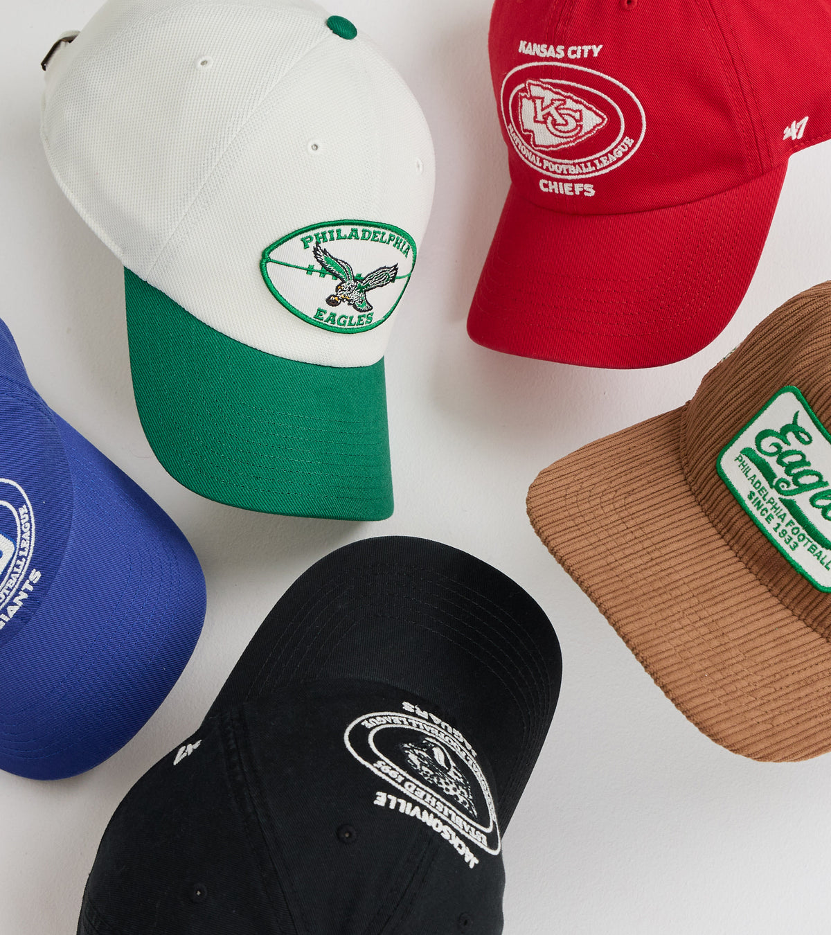 Five baseball caps arranged on a white background: a white and green Philadelphia Eagles cap, a red Kansas City Chiefs cap, a blue cap partially visible at left, a black cap at bottom center, and a brown corduroy cap at right with a green embroidered patch.