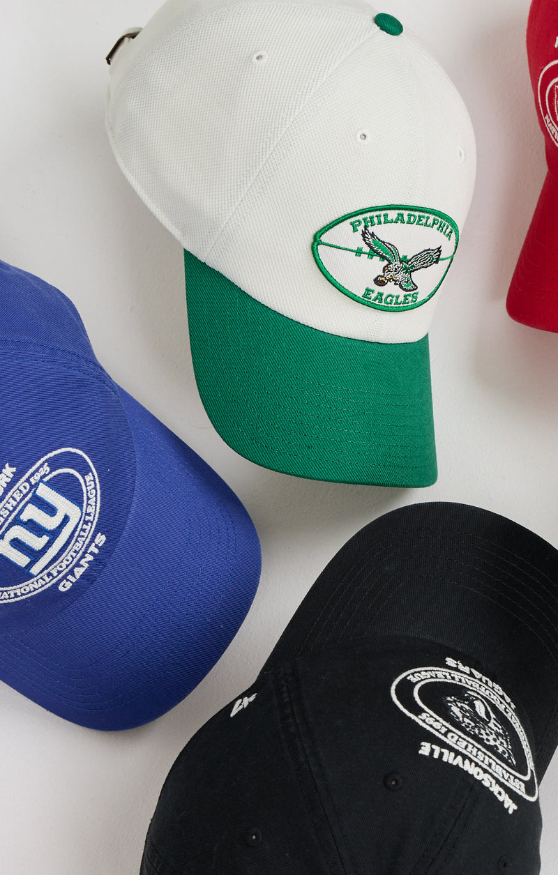 A set of baseball caps arranged on a white surface, including a Philadelphia Eagles cap with a white crown and green brim, a blue New York Giants cap, a black cap with a white logo, and a partial red cap at the edge.
