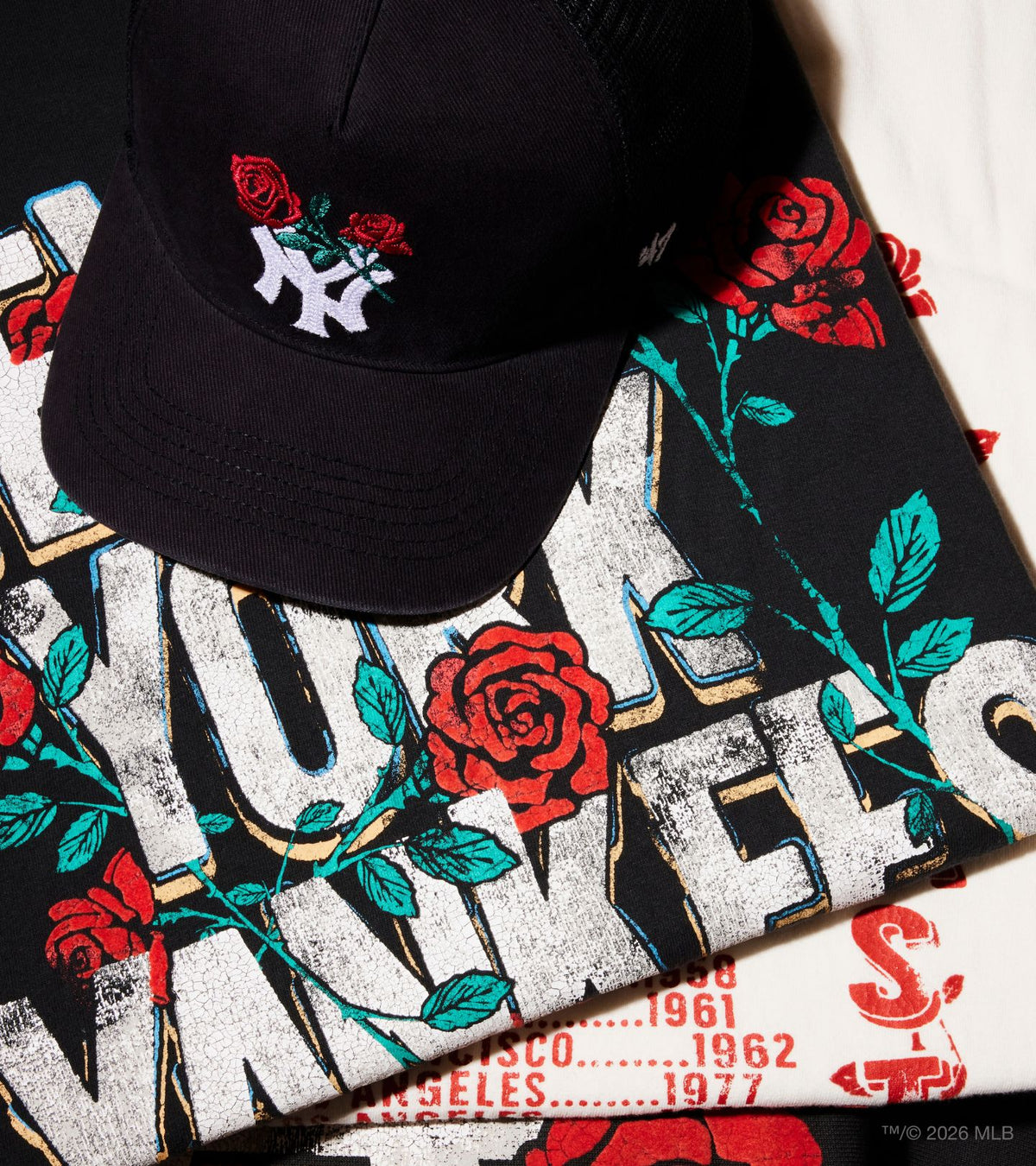 A black baseball cap with a white and red embroidered New York Yankees logo and red roses, resting on top of a black graphic T-shirt featuring distressed white lettering, red roses, and teal leaves.